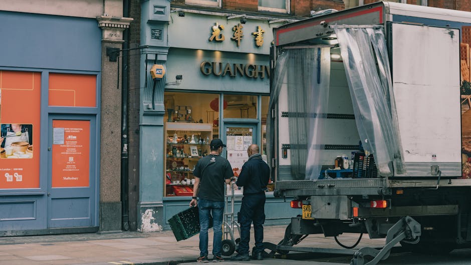 Two movers from Harringay Removals, dressed in dark clothing, are preparing to load a truck outside a shop with a sign that reads 'Guang.' One mover is lifting a black plastic crate, while the other i