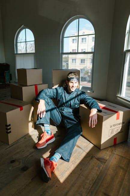 A person dressed in a blue jumpsuit, sitting on the wooden floor of a bright room with large arched and rectangular windows, surrounded by several closed cardboard boxes sealed with red and black tape