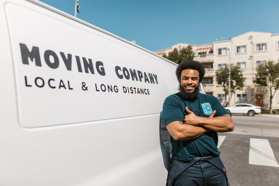 A smiling man with a curly afro hairstyle and a full beard stands next to a white moving van parked on a street in an urban area with modern residential buildings in the background. He is wearing a da
