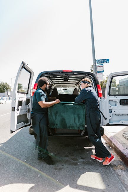 Two movers, wearing dark uniforms, are carefully loading a large, green-shaped upholstered piece of furniture into the back of a white cargo van parked on a paved street. The van’s rear doors are fu