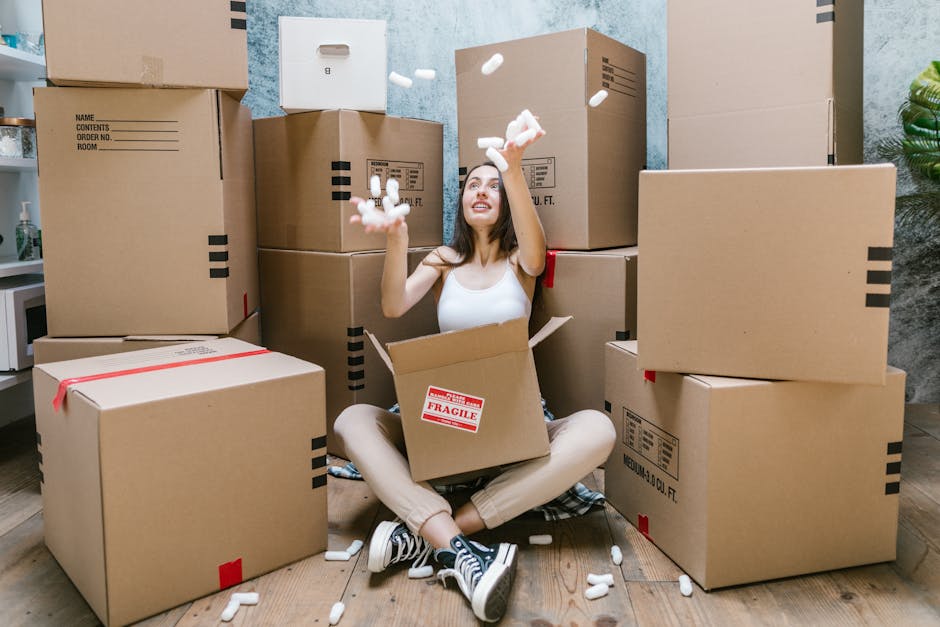 A man with dark curly hair and a beard, wearing a blue t-shirt, is inside a room with a wooden ceiling and white walls, standing behind several large cardboard moving boxes stacked at waist height. Th