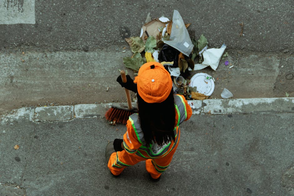 A person dressed in high-visibility orange work clothing and a matching cap is standing on a pavement next to a curb, holding a large outdoor broom with a wooden handle and red bristles, during a home