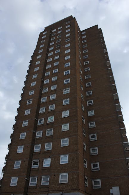 A tall multi-storey block of flats constructed from reddish-brown bricks, with multiple white-framed windows arranged in a grid pattern, extending upward into a cloudy sky. The building's exterior app