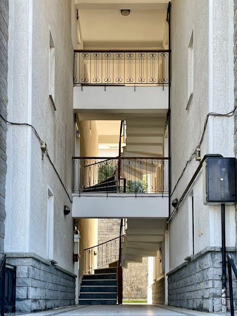 The image depicts a narrow, multi-story staircase of an apartment building with three visible floors, each featuring a small balcony with decorative black wrought iron railings. The exterior walls are
