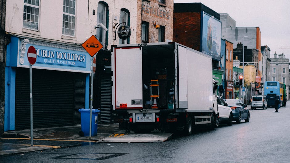 A white box truck with its rear doors open is parked on a city street outside a building with a blue sign reading 'Dublin Mouldings' and a Guinness logo, indicating a premises related to plaster work.