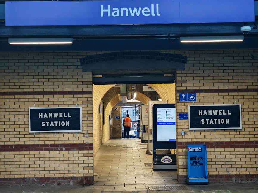 Image of the exterior entrance to Hanwell Station, displaying brickwork with two black and white signs reading 'HANWELL STATION' mounted on either side of a doorway. Above the entrance, a blue sign wi